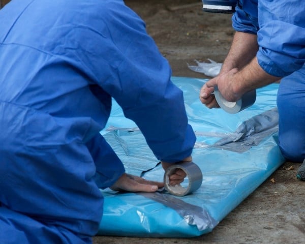 Two workers in blue uniforms installing or sealing a blue plastic sheet or tarp on the ground